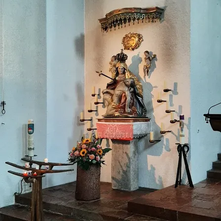 Eine Statue einer gekrönten Frau, die ein Kind hält, steht auf einem Podest mit Kerzen. Unten steht eine Blumenvase. Die Wand hat einen goldenen Zierrat und ein rotes Schild.