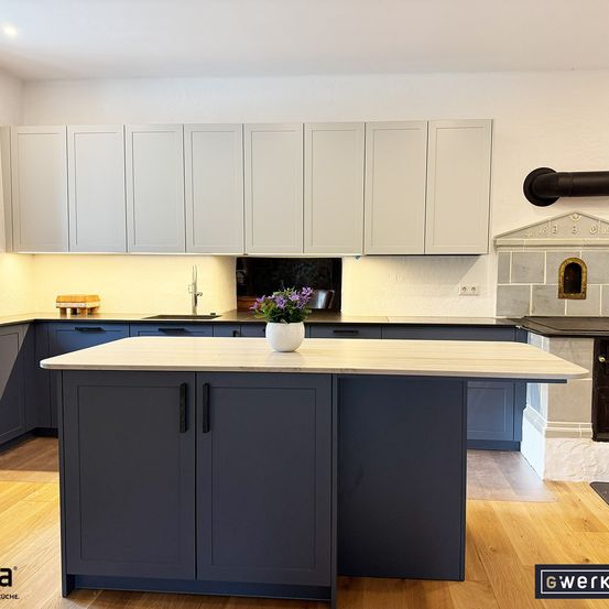 Eine moderne Küche mit weißen Schränken, einer dunkelblauen Insel, einer weißen Arbeitsplatte und einer weißen Vase mit lila Blumen darauf. Es gibt einen Holzboden und eine weiße Decke.