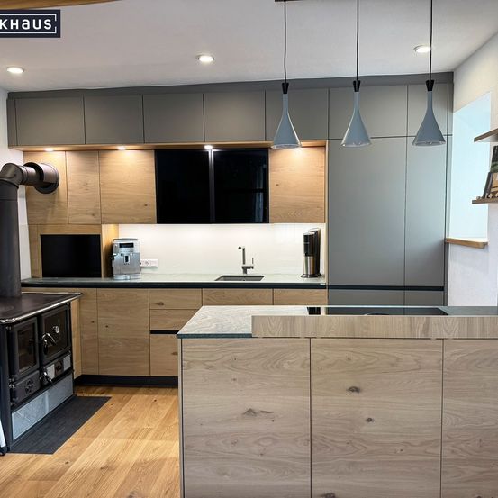 Eine moderne Küche mit Holzschränken, einer zentralen Insel und einem Ofen. Über der Arbeitsplatte hängen drei Pendelleuchten.