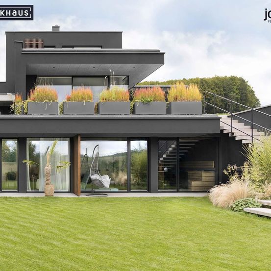 Modernes schwarzes Haus mit einem Balkon, der große rechteckige Pflanzgefäße mit hohen, goldfarbenen Pflanzen aufweist. Das Haus hat eine große Glaswand, eine Glastür und eine Hängeschaukel. Die Landschaft umfasst einen Rasen, einige Pflanzen und eine Treppe.