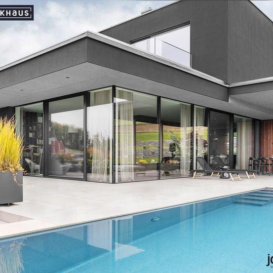 Ein modernes Haus mit Glaswänden und einer großen Terrasse, mit Swimmingpool, Liegestühlen und einem Holzpfeiler.