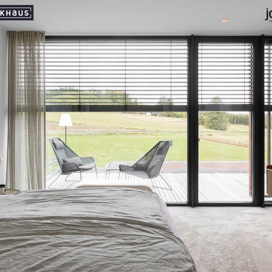 Modernes Schlafzimmer mit großen Fenstern, großem Bett und Außenterrasse mit Stühlen und einer Lampe. Blick auf eine Wiese durch die Fenster.