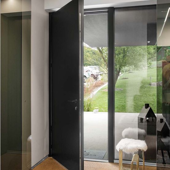 Eine offene schwarze Tür führt zu einem Blick auf einen Rasen mit Bäumen und geparkten Autos draußen. Eine Glaswand spiegelt einen Hocker und ein hausartiges Gebäude wider.