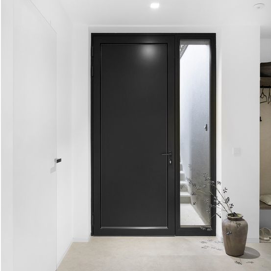 Ein minimalistischer Innenraum mit einer großen schwarzen Tür mit Glasfenster und silberner Klinke. Ein Topf mit einer Pflanze steht auf dem Boden. Der Raum hat weiße Wände und einen Holzboden.