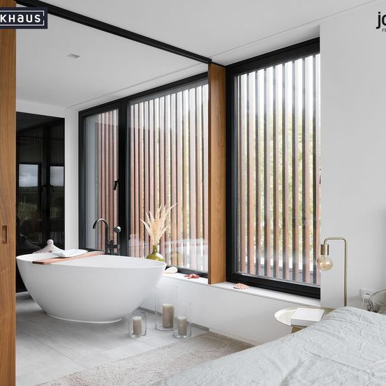 Ein modernes Badezimmer mit einer weißen Badewanne und einem großen Fenster mit Holzjalousien. Der Raum verfügt über weiße Wände, Holzböden und einen Nachttisch mit einer Lampe.