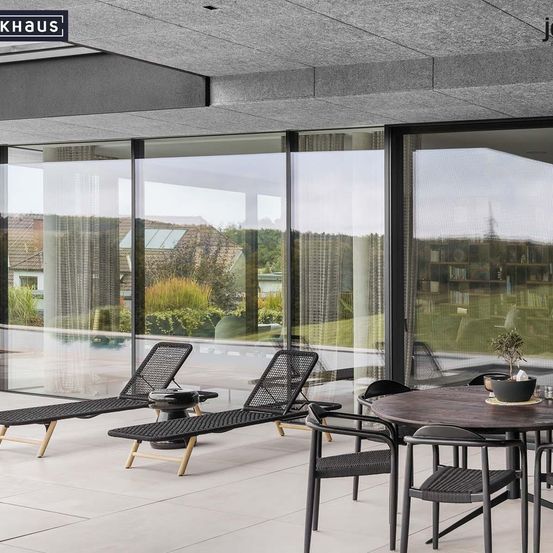 Eine Außenterrasse mit Liegestühlen, einem Tisch und großen Glasfenstern, die einen Blick auf einen Pool und einen Garten bieten. Die Terrasse ist von Grünpflanzen umgeben und ein Haus im Hintergrund.