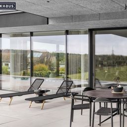 Eine Außenterrasse mit Liegestühlen, einem Tisch und großen Glasfenstern, die einen Blick auf einen Pool und einen Garten bieten. Die Terrasse ist von Grünpflanzen umgeben und ein Haus im Hintergrund.