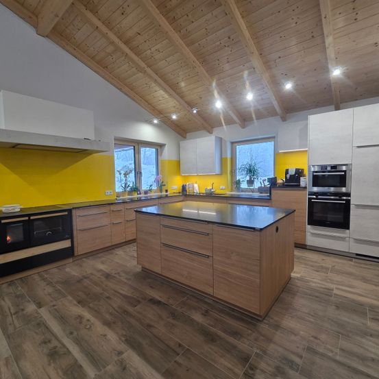 Eine moderne Küche mit Holzschränken, einer gelben Wand und einer Holzdecke. Die Küche hat eine zentrale Insel, eine Kaffeemaschine und einen Herd. Der Boden ist aus Holz.