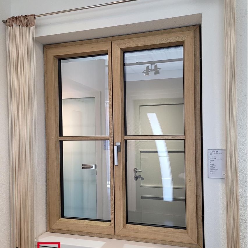 Ein hölzerne Doppelfenster mit Glasscheibe und silbernen Griffen. Das Fenster spiegelt den Innenraum und eine an der Decke montierte Lampe wider.