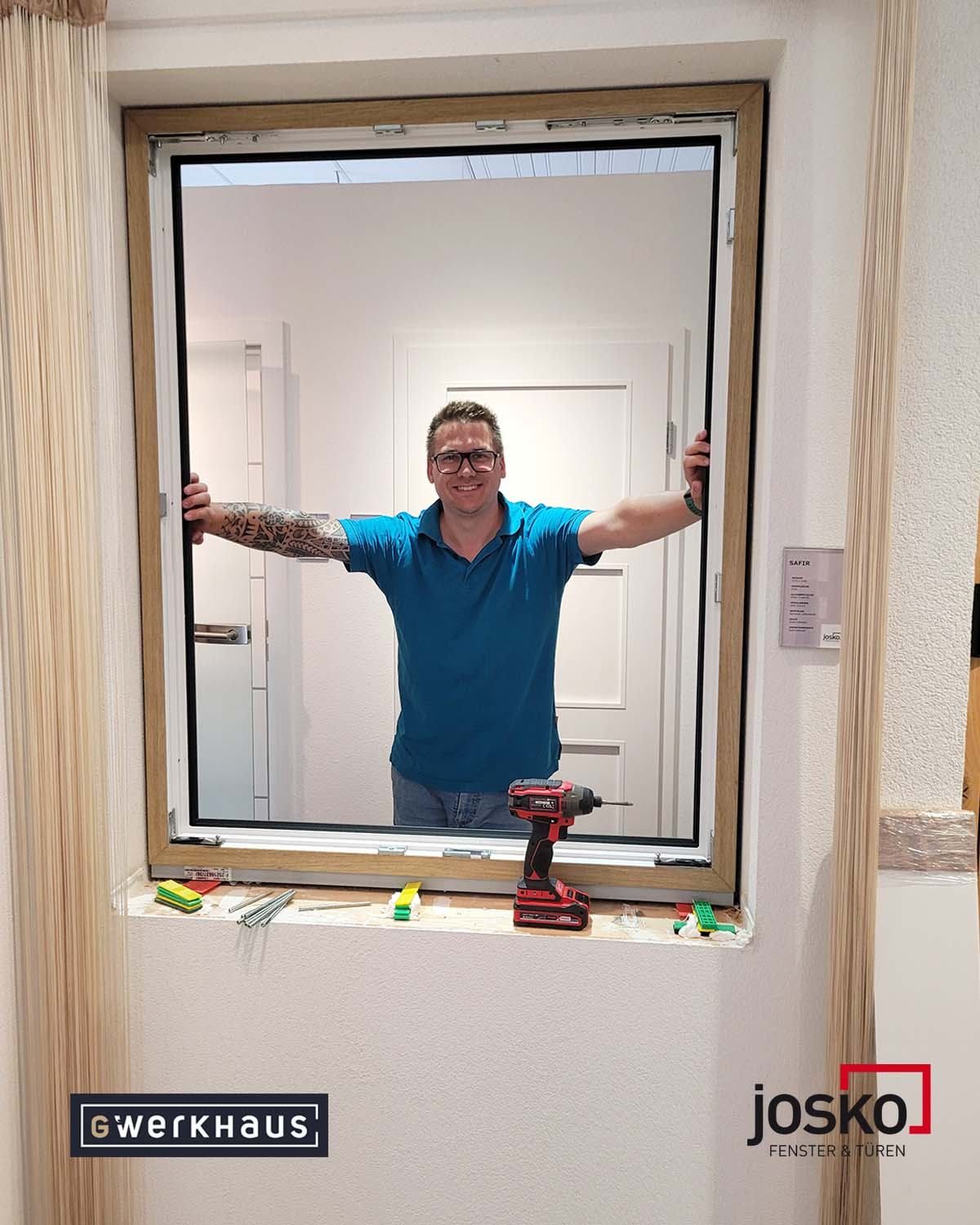 Ein Mann mit Tätowierungen an den Armen lächelt und lehnt sich an eine Tür, hält eine Bohrmaschine. Werkzeuge liegen auf der Fensterbank.