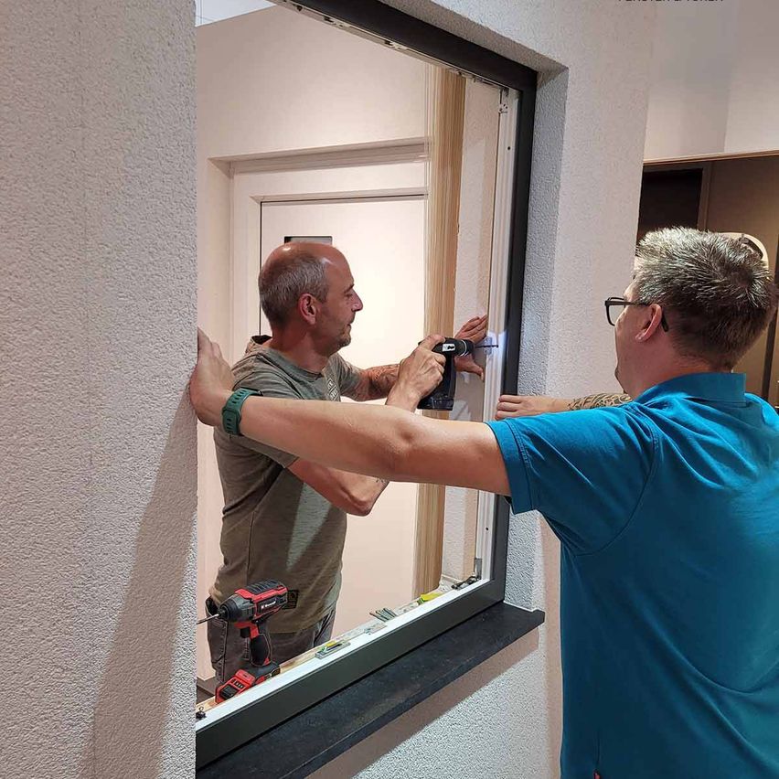 Zwei Männer arbeiten an einem Fensterrahmen. Einer bohrt, während der andere den Rahmen hält. Sie tragen beide Freizeitkleidung.