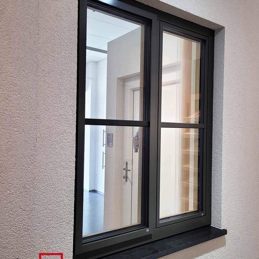 Ein schwarz gerahmtes Fenster mit klaren Glasscheiben, montiert an einer strukturierten weißen Wand, reflektiert einen Raum mit einer weißen Tür und einem silbernen Griff.