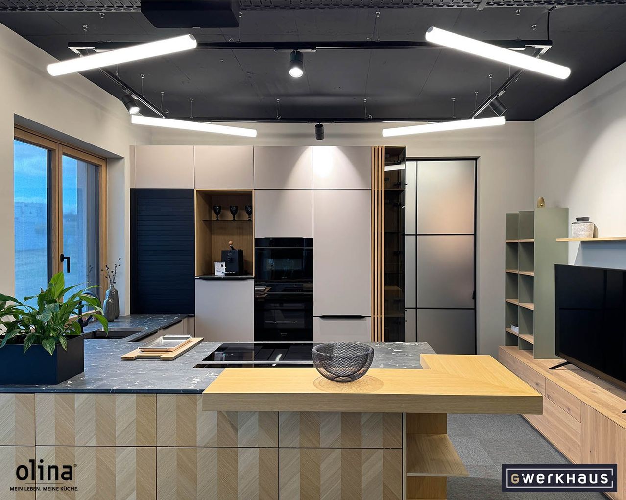 A modern kitchen with gray and white cabinets, a black stove, and wooden countertops. The ceiling features linear lighting. A basket sits on the counter, and a TV is on the right.