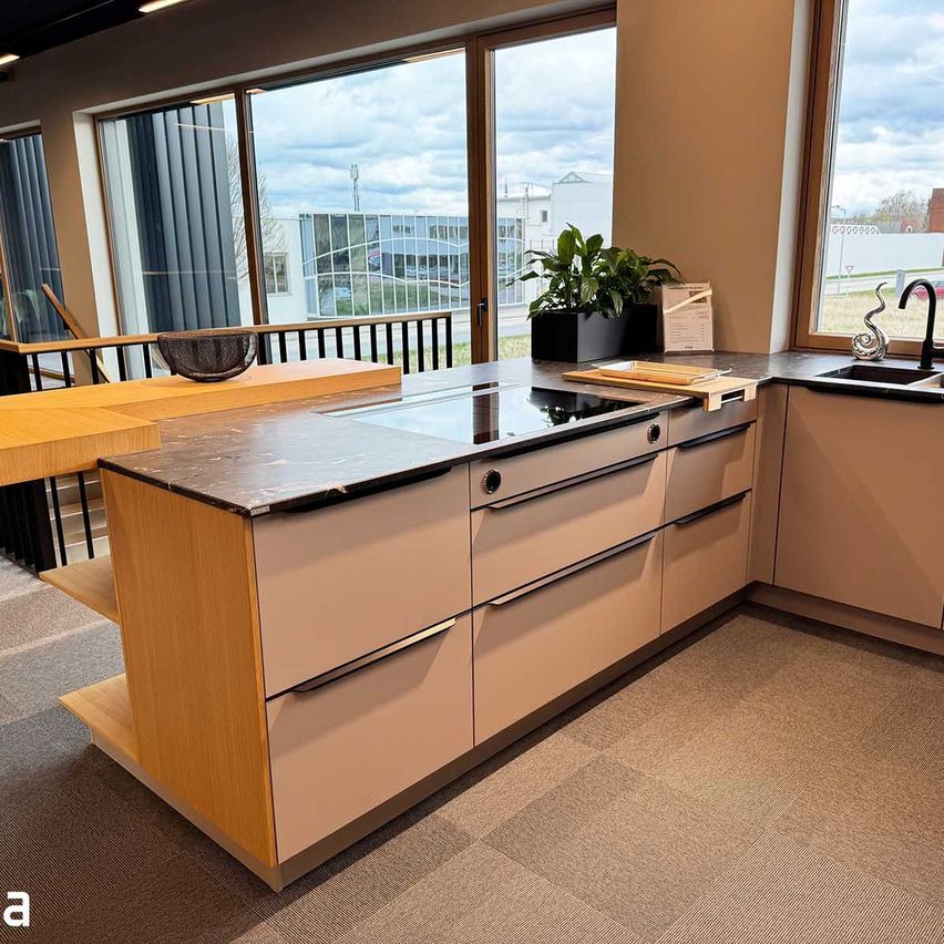 A modern kitchen with a marble countertop, a black stove, and a sink with a faucet. There is a potted plant and a basket on the countertop. The kitchen has large windows and a balcony.