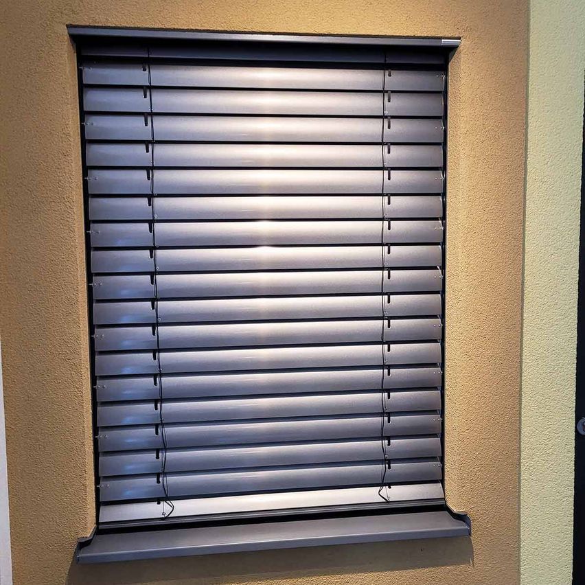 Nahaufnahme eines Fensters mit geschlossenen silbernen horizontalen Jalousien an einer beigen Wand.