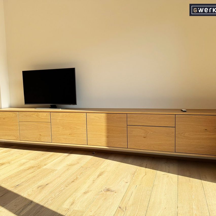 A modern wooden entertainment center with a flat-screen TV on top and a remote control on the right side, placed in a room with a beige wall and wooden floor.