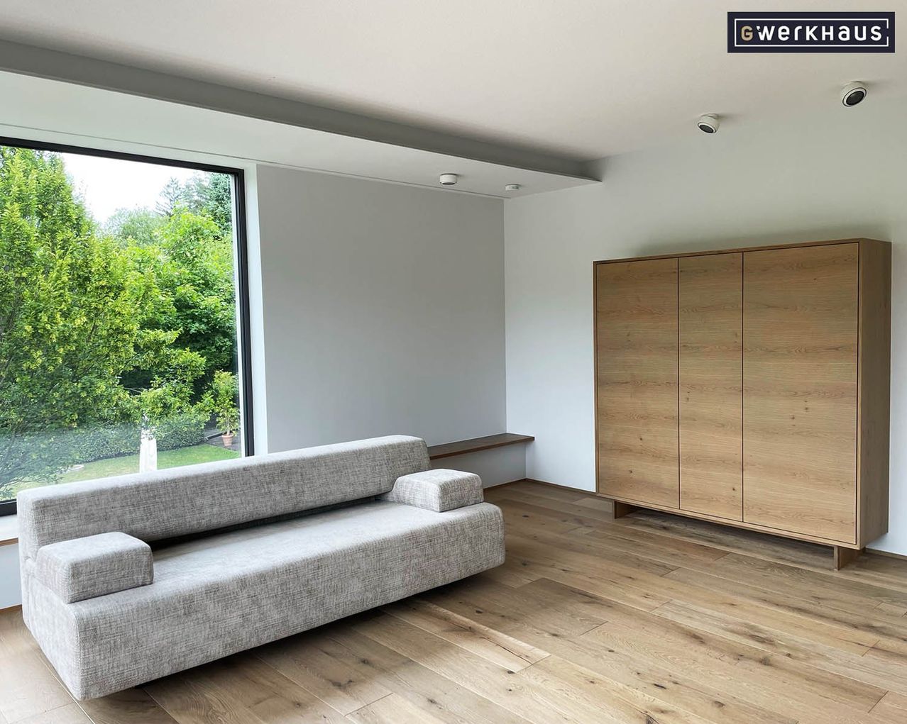 An empty living room with a gray sofa, wooden floor, and a large window overlooking a garden.