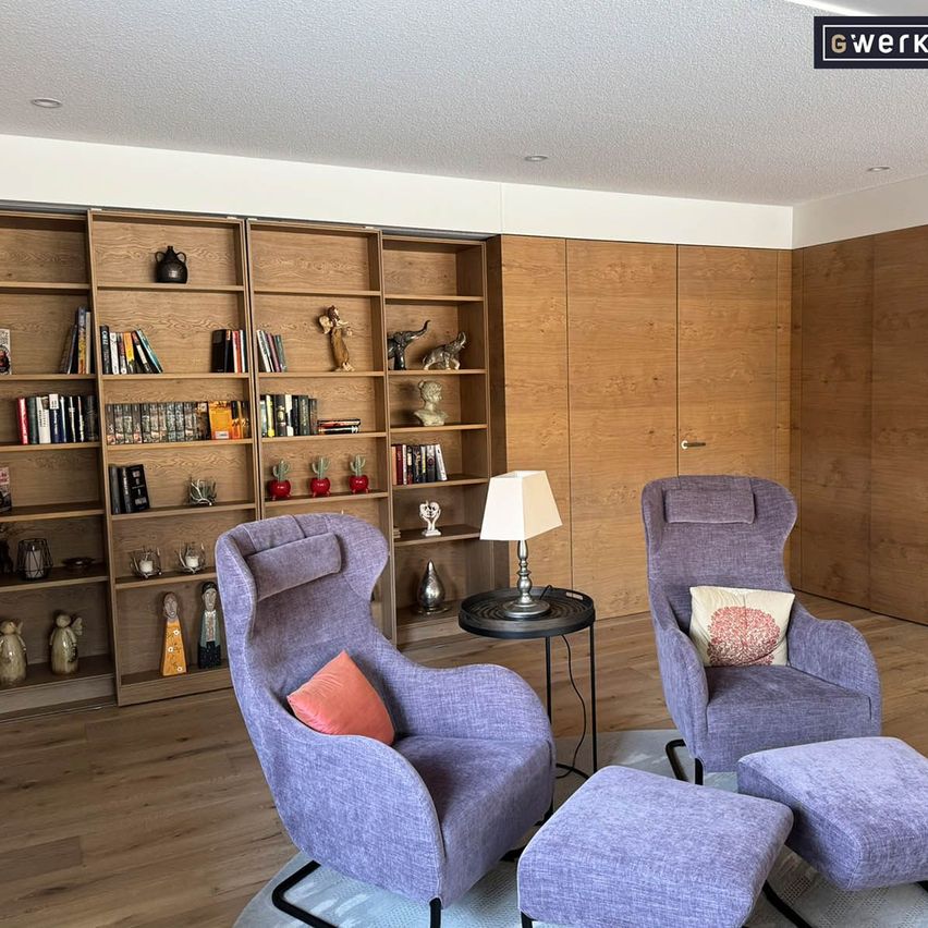 A cozy living room with two purple armchairs, a round table with a lamp, and a wooden bookshelf filled with books and decorative items.