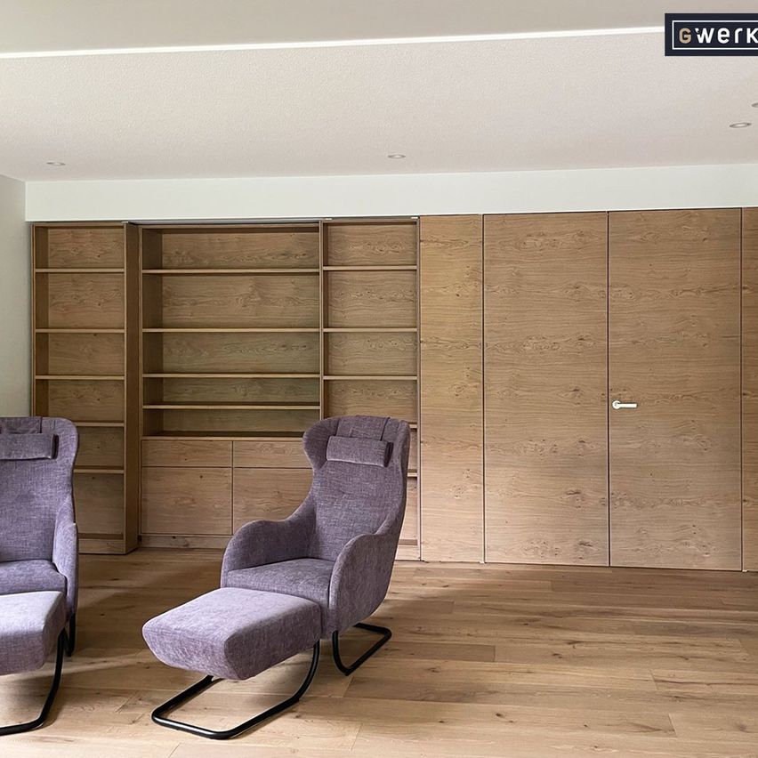 A modern living room with a wooden floor, two gray armchairs, and a wooden wall with shelves and cabinets.