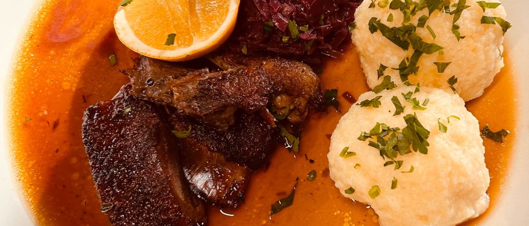 Ein Teller mit geschmortem Rindfleisch, Rotkohl und Kartoffelknödel, garniert mit einer Zitronenscheibe und Petersilie.