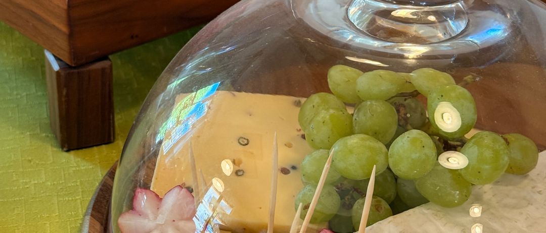 Ein Holztablett mit einer Glasglocke hält eine Auswahl an Käse, Trauben und Radieschen. Daneben befindet sich ein Tablett mit geschnittenem Fleisch, ebenfalls unter einer Glasglocke.