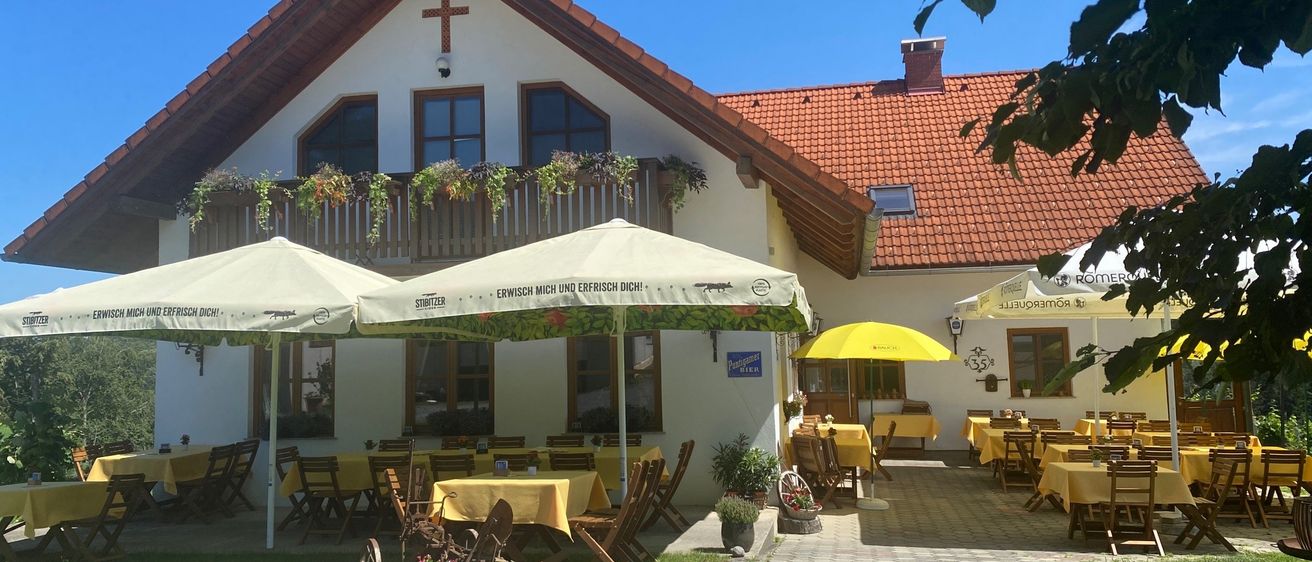 Ein weißes Haus mit rotem Dach, einem Kreuz oben und einem Balkon mit Topfpflanzen. Es hat mehrere Fenster und eine offene Terrasse mit gelben Tischdecken, Stühlen und Sonnenschirmen.