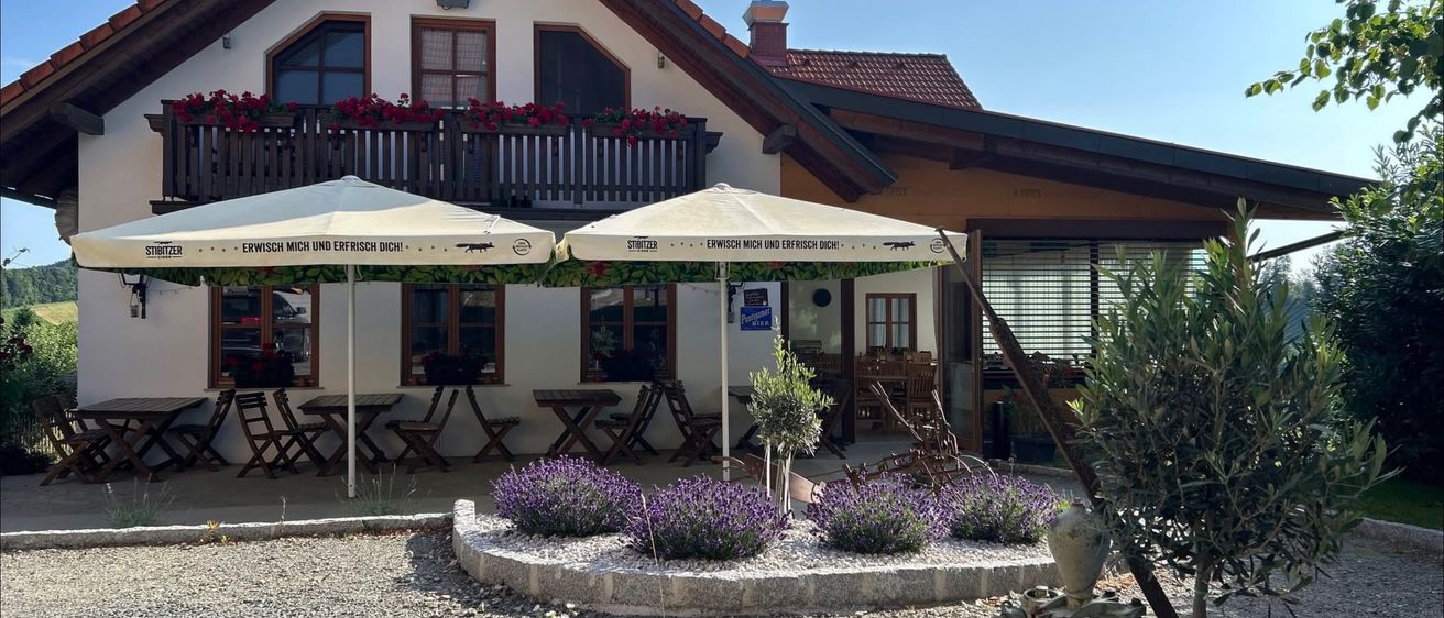 Ein charmantes Haus mit einem Balkon voller roter Blumen und einem Kreuz auf dem Dach. Die Vorderseite hat zwei Schirme und einen kleinen Garten mit lila Blumen.
