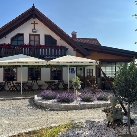 Ein Haus mit einem Kreuz auf seinem Giebel verfügt über zwei Außenschirme. Rote Blumen befinden sich auf dem Balkon. Der Eingang hat Glastüren. Ein Garten mit Lavendelpflanzen steht davor.