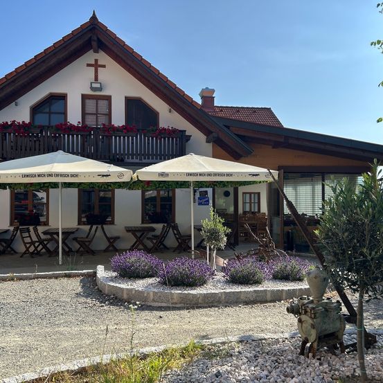 Ein Haus mit einem Kreuz auf seinem Giebel verfügt über zwei Außenschirme. Rote Blumen befinden sich auf dem Balkon. Der Eingang hat Glastüren. Ein Garten mit Lavendelpflanzen steht davor.