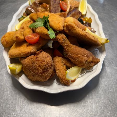 Ein weißer Teller mit gebratenem Hühnchen, paniertem Fleisch, Gemüse und Zitronenspalten. Garniert mit Petersilie und Tomaten.