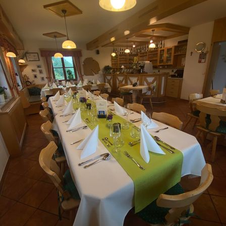 Ein Esszimmer mit einem langen Tisch, der mit weißer und grüner Tischdecke bedeckt ist, mit Weinglas, Besteck und Servietten bestückt. Der Raum hat Holzstühle, Lampen und eine Bartheken.