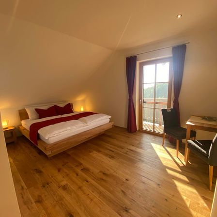 Ein gemütliches Schlafzimmer mit Holzboden, einem Bett, roten Kissen und Lampen. Ein Tisch und Stühle stehen in der Nähe einer Glastür mit Vorhängen.