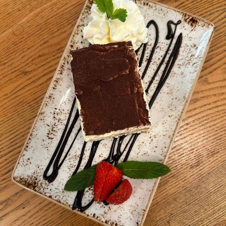 Ein Schokoladen-Tiramisu-Dessert auf einem weißen Teller, garniert mit Schlagsahne und Schokoladensauce, verziert mit Erdbeeren und Minzblättern auf einem Holztisch.