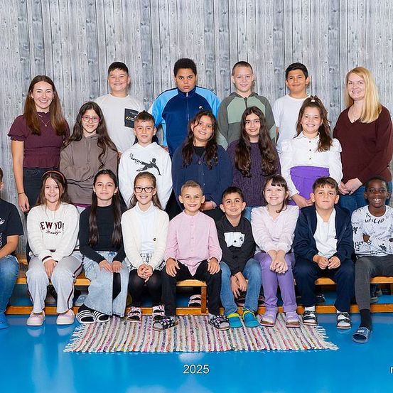 Gruppenfoto von Schülern und Lehrern in einer Turnhalle, datiert auf 2025, mit hölzernem Hintergrund und blauem Boden.