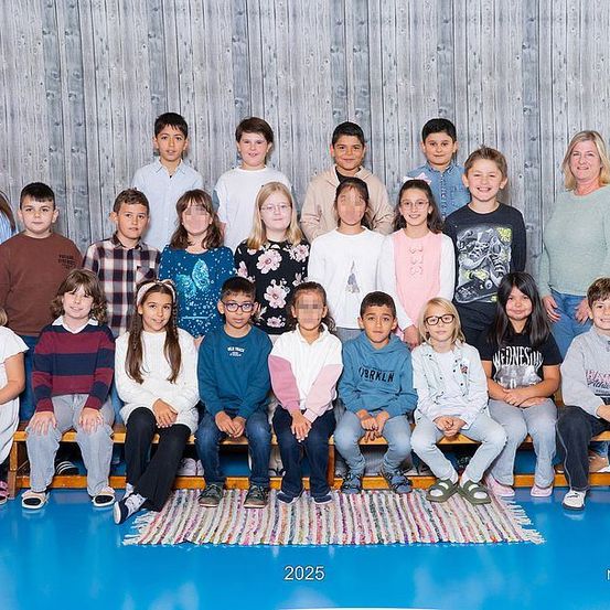 Eine Gruppenaufnahme von Schülern und Lehrern in einem Klassenzimmer, sitzend auf einer Bank und stehend vor einer Holzwand, datiert auf 2025.