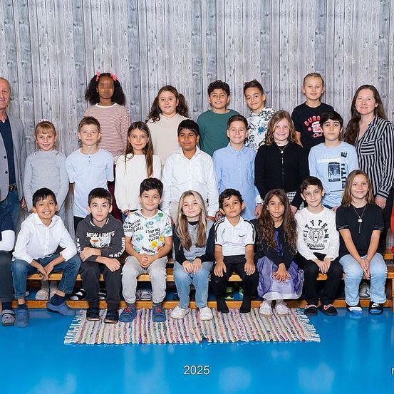 Eine Gruppenaufnahme von Schülern und Lehrern in einer Turnhalle, aufgenommen im Jahr 2025. Die Schüler sitzen und stehen auf Bänken und tragen verschiedene Kleidungsstücke. Im Hintergrund befindet sich eine Holzwand und ein Teppich.