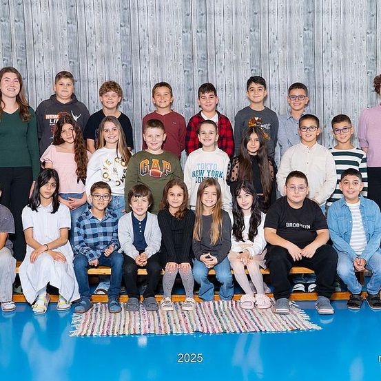 Ein Klassenfoto mit einer Gruppe von Schülern und zwei Lehrerinnen, aufgenommen im Jahr 2025, vor einer Holzwand. Die Schüler sitzen auf einer Holzbank, während einige stehen. Ein Teppich liegt auf dem blauen Boden.