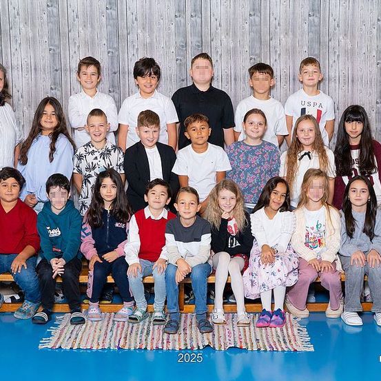 Eine Gruppe von Kindern und zwei Erwachsenen posiert für ein Foto vor einer hölzernen Kulisse. Die Kinder sitzen auf einer Bank, einige stehen. Sie tragen alle Freizeitkleidung. Das Bild hat einen Wasserzeichen mit dem Text 2025 in der unteren Mitte.