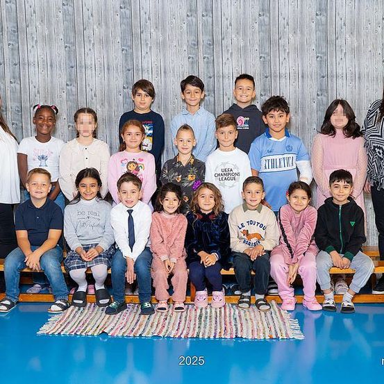 Eine Gruppenaufnahme von Schülern und Lehrern, die stehend und sitzend auf einer Bank mit einem Teppich davor. Das Foto wurde in einem Raum mit einer Holzwand aufgenommen.