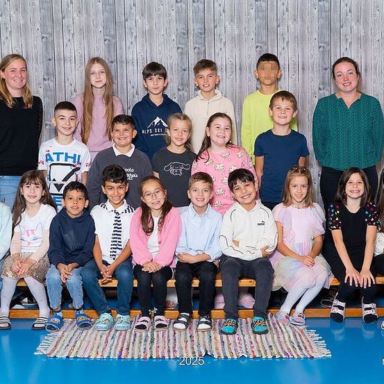 Eine Gruppe von Kindern und zwei Frauen sitzen zusammen auf einer Bank in einem Raum. Sie lächeln und posieren für ein Foto. Ein Teppich liegt auf dem Boden. Der Wasserzeichen befindet sich in der unteren rechten Ecke.