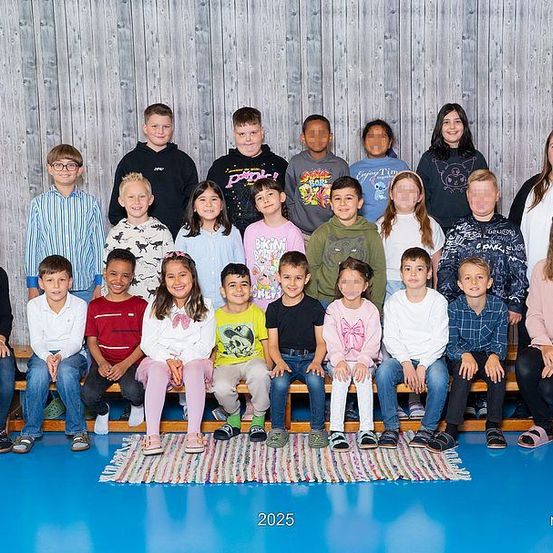 Eine Gruppe von Kindern und Lehrern steht vor einer Holzwand, trägt Freizeitkleidung, einige mit Brille. Die Kinder posieren für ein Foto.