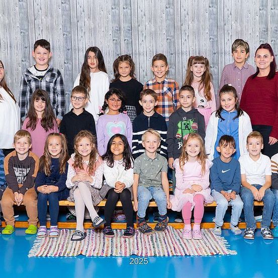 Eine Gruppenaufnahme von Kindern und Erwachsenen, einschließlich Lehrern, in einem Raum mit blauem Boden und einer Holzwand. Die Kinder sitzen auf einer Bank, während andere hinter ihnen stehen. Das Foto ist mit dem Jahr 2025 beschriftet.