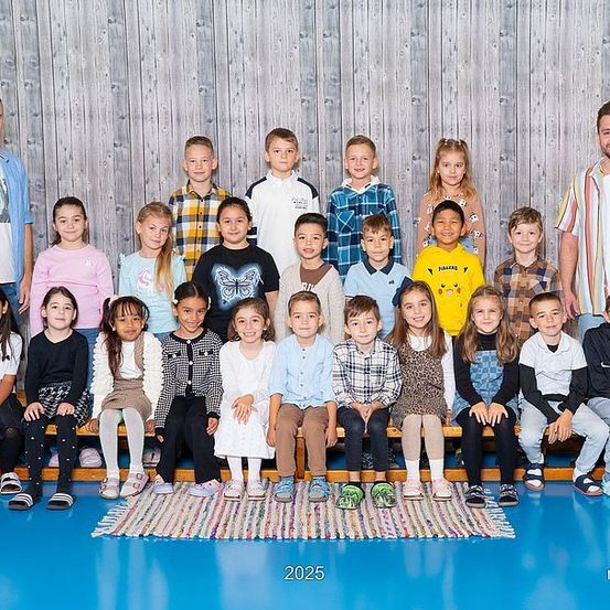Ein Klassenfoto von Schülern und zwei Lehrern, einem Mann und einer Frau, die auf einer Bank in einer Turnhalle sitzen. Das Jahr 2025 steht auf dem Boden geschrieben.