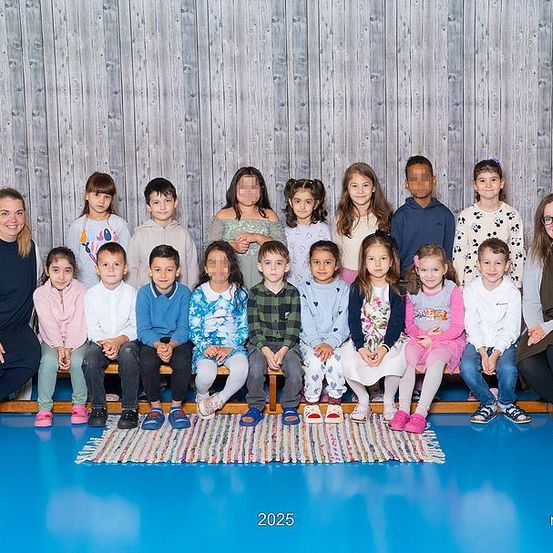 Klassenfoto aus dem Jahr 2025 mit Schülern und zwei Lehrern, mit verpixelten Gesichtern. Die Umgebung ist drinnen, vor einer Holzwand, mit blauem Boden und einem Teppich.
