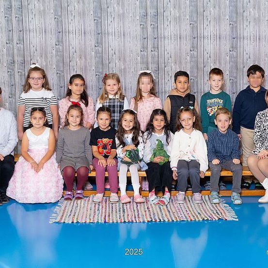 Ein Gruppenfoto von jungen Kindern und ihren beiden Lehrerinnen. Sie sitzen alle auf einer Bank vor einer Holzwand und tragen verschiedene Kleidungsstücke. Das Bild ist auf das Jahr 2025 datiert.