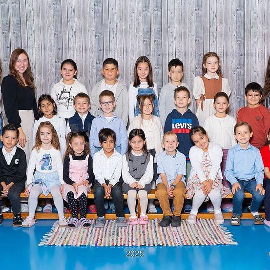 Eine Gruppe von Kindern und Erwachsenen posiert für ein Foto in einer Turnhalle. Die Kinder sitzen auf einer Bank, während die Erwachsenen hinter ihnen stehen. Das Bild hat unten einen Wasserzeichen.
