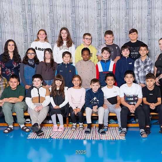 Eine Gruppe von Schülern und Lehrern posiert für ein Foto auf einem blauen Boden in einem Raum mit einer Holzwand. Die Schüler sitzen auf einer Bank.