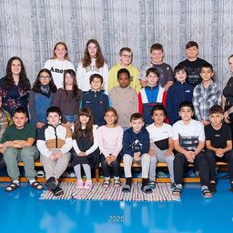 Eine Gruppe von Schülern und Lehrern posiert für ein Foto auf einem blauen Boden in einem Raum mit einer Holzwand. Die Schüler sitzen auf einer Bank.