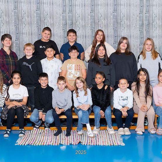Ein Gruppenfoto von Schülern in einem Klassenzimmer. Sie sitzen auf einer Holzbank und stehen gegen eine Holzwand. Sie alle lächeln und posieren für das Foto. Der Boden ist mit einem blauen Teppich bedeckt. Das Bild hat einen Wasserzeichen in der unteren rechten Ecke.