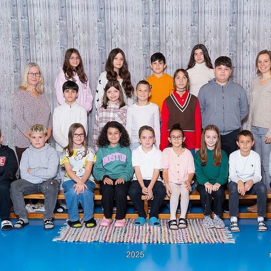 Eine Gruppe von Kindern und einigen Erwachsenen posiert für ein Foto in einem Studio mit einer Holzwand. Die Kinder sitzen auf Bänken, während einige Erwachsene hinter ihnen stehen. Der Boden ist mit einem Teppich bedeckt. Das Foto hat einen Wasserzeichen in der unteren rechten Ecke.